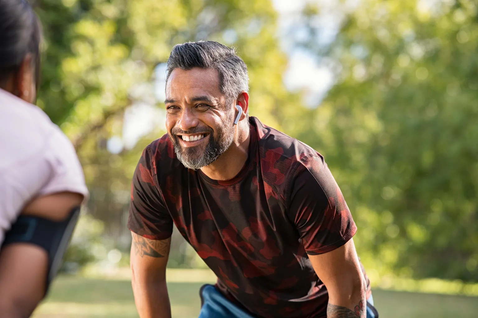 A smiling man in his forties with a grey beard resting his hands on his knees after a run in the park