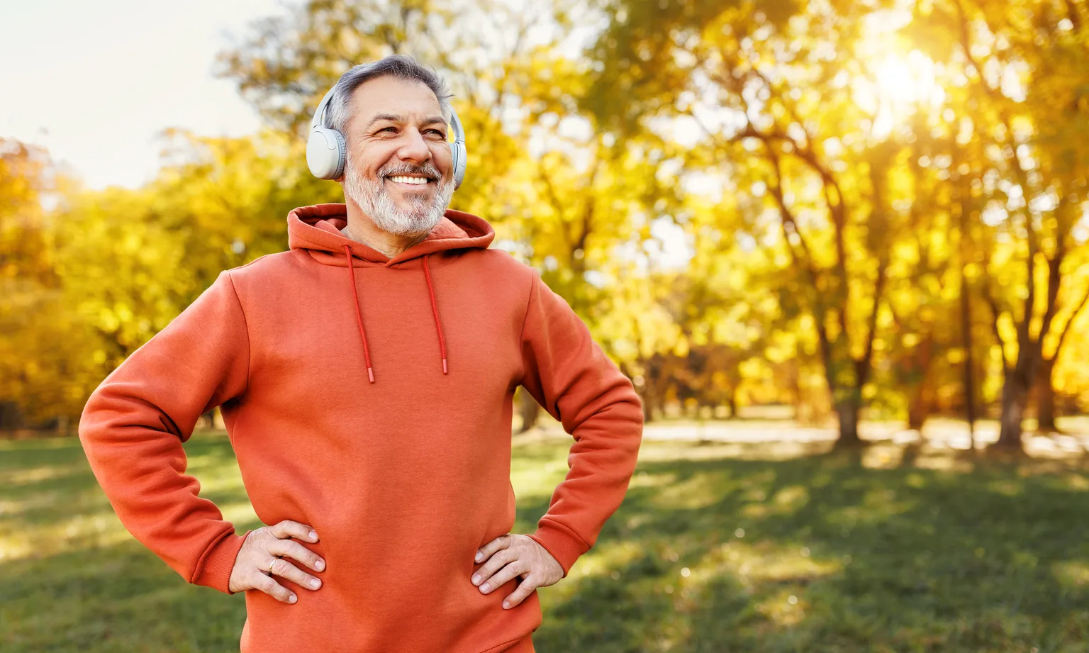 A smiling man in his fifties in a red hoodie and headphones standing in a sunny park after a workout