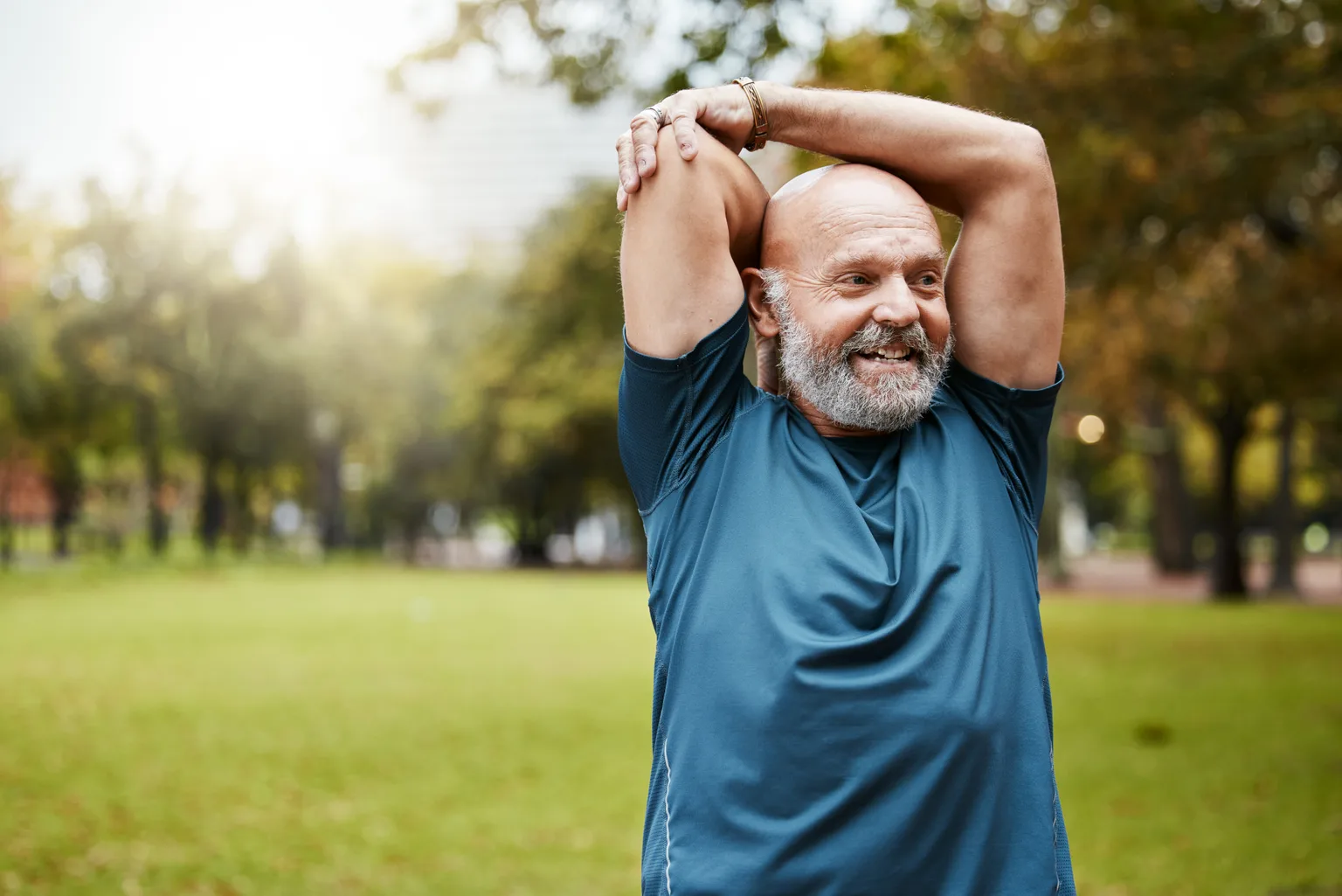 A fit older man in a blue T-shirt stretching his shoulder and triceps in a sunlit park