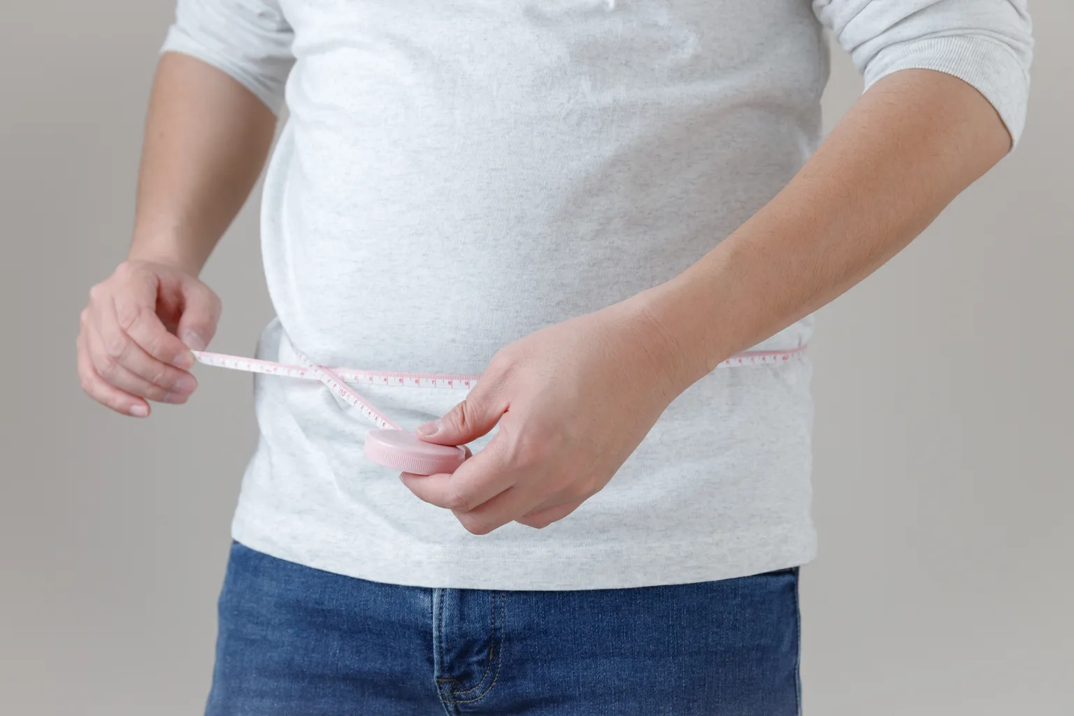 A man in a grey T-shirt and jeans measuring his waist with a tape measure