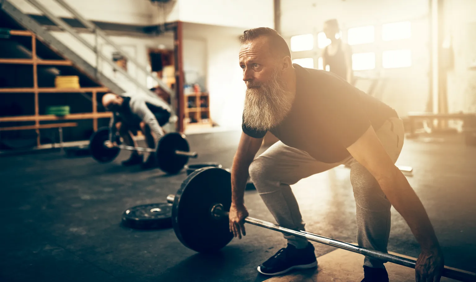 A bearded man in his fifties setting up to deadlift a barbell in a dark gym