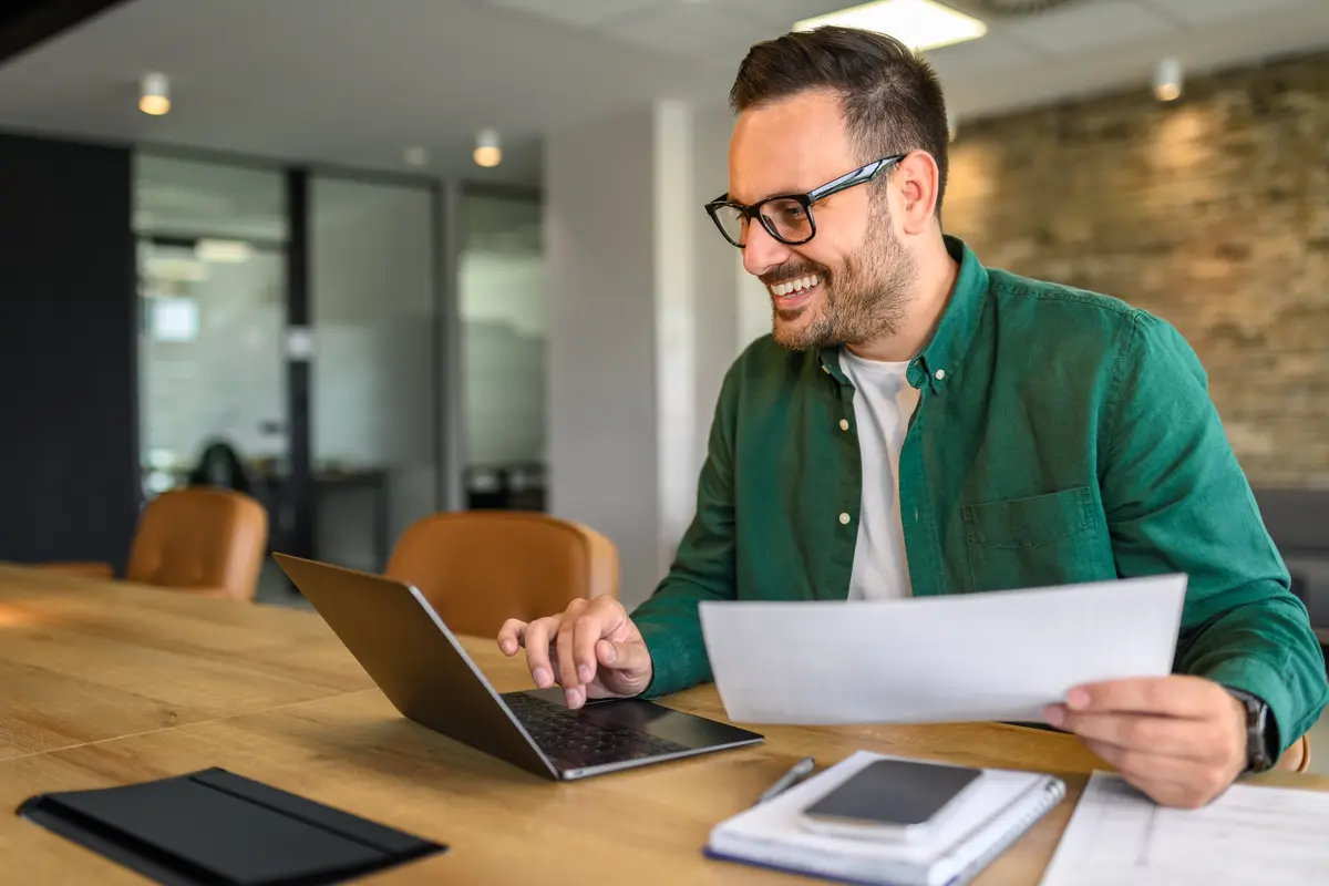 Smiling man reviewing a paper at his laptop